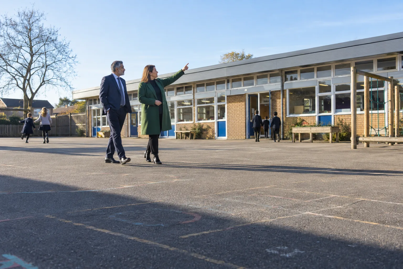 A trust's finance director and a primary headteacher cross an academy playground on a bright morning, the headteacher pointing up at a newly replaced roof