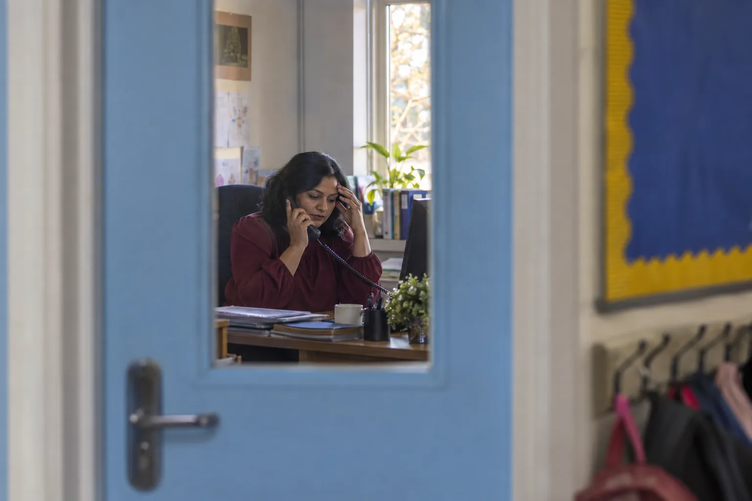 Seen through the glass panel of her closed office door, a headteacher speaks on the phone at her desk, the corridor softly out of focus in the foreground