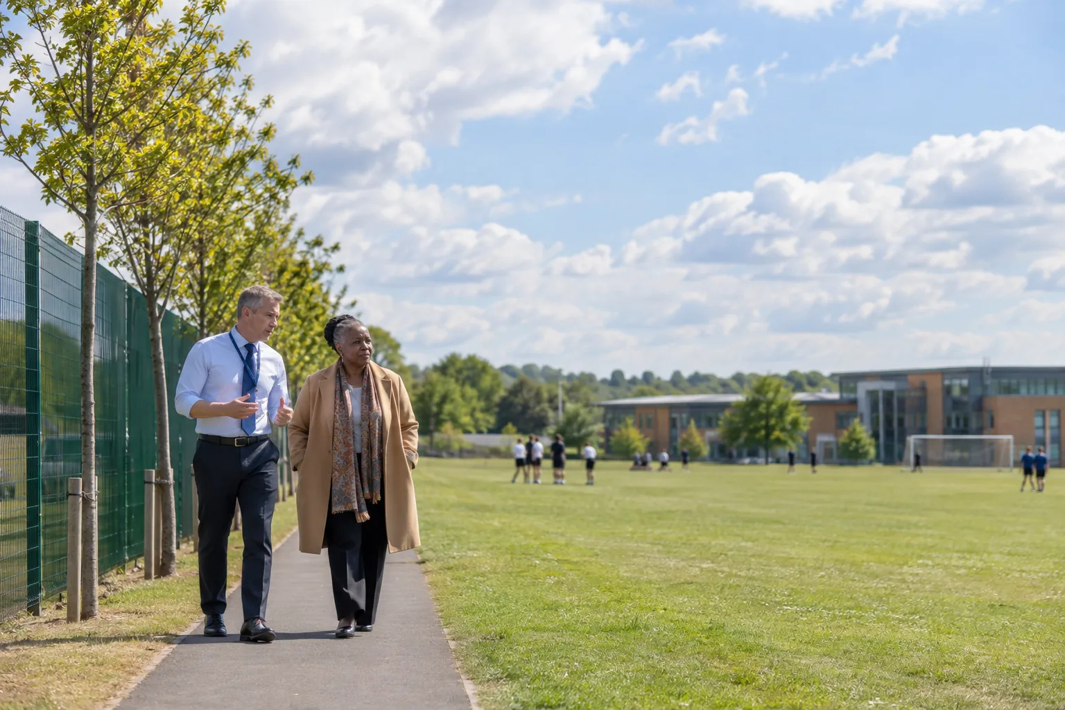 A headteacher and the chair of governors walk the perimeter path of a school playing field at midday, deep in conversation