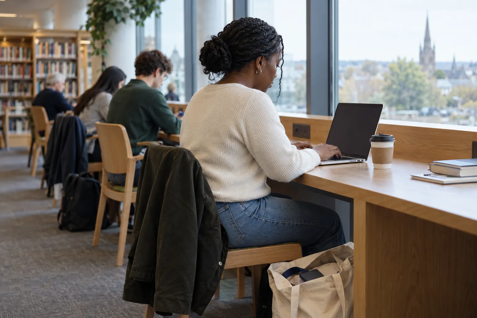 A charity finance officer works on her own laptop at a study desk in a public library on a Saturday morning, a work lanyard tucked into the open bag at her feet