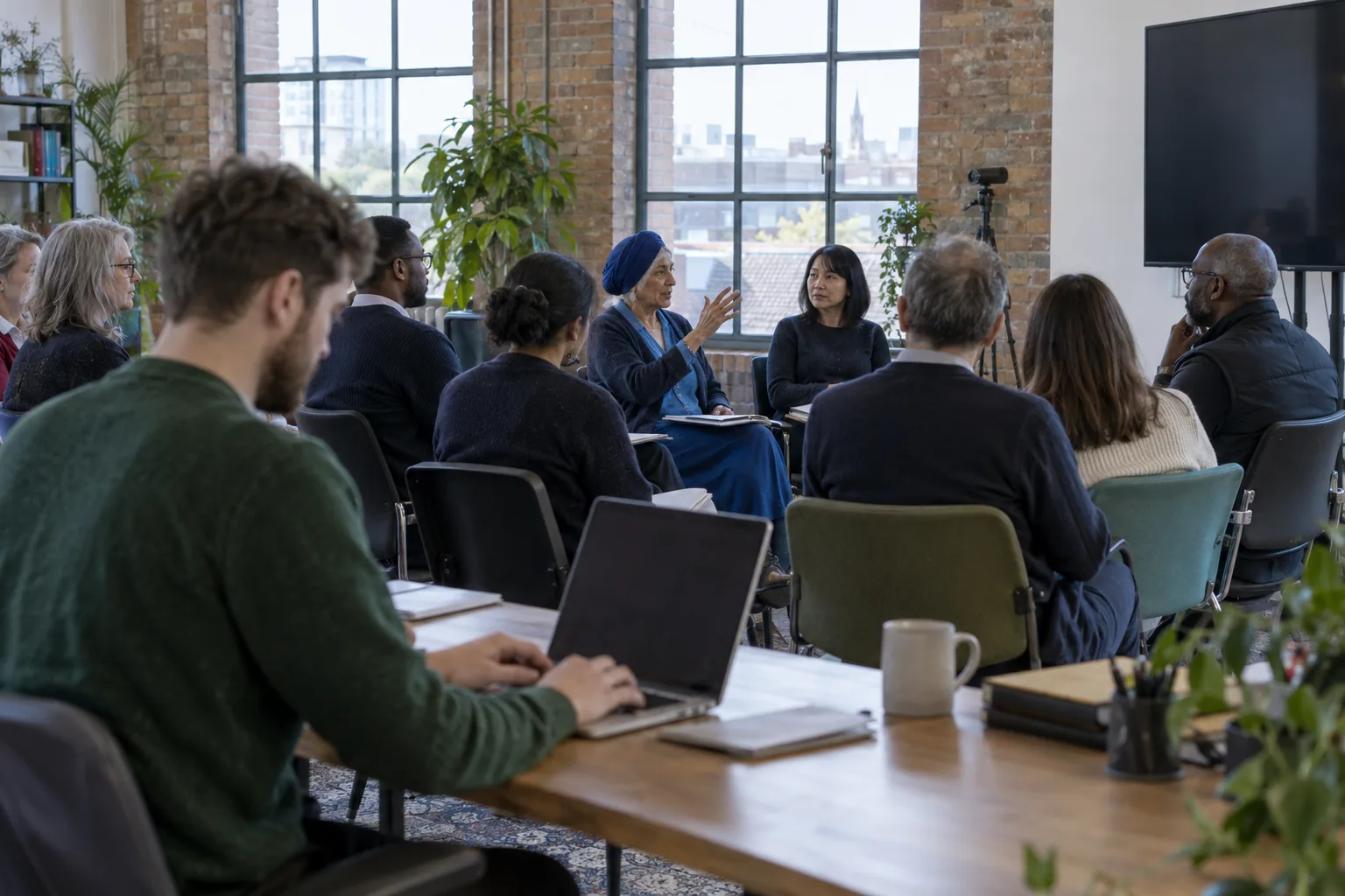 At a hybrid annual general meeting in a small charity’s brick-walled office, a member speaks from her seat while the minute-taker types at a side desk and a webcam on a tripod faces the room
