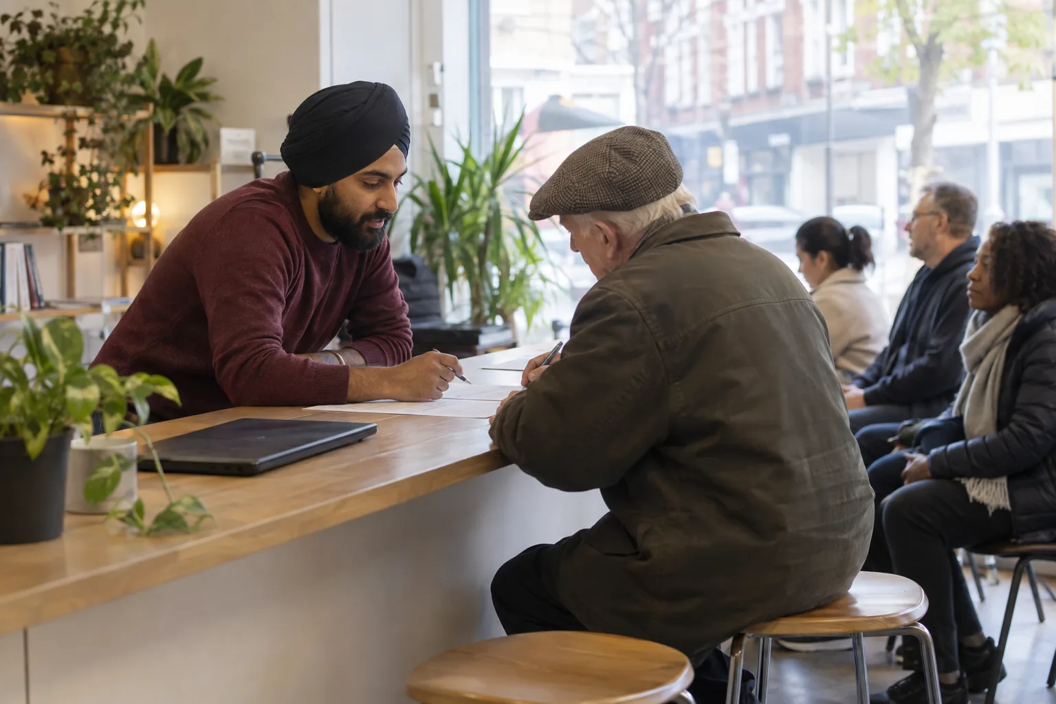 A volunteer at a community advice charity helps an older man complete a paper form at a drop-in counter in a busy high-street shopfront, morning light through the window