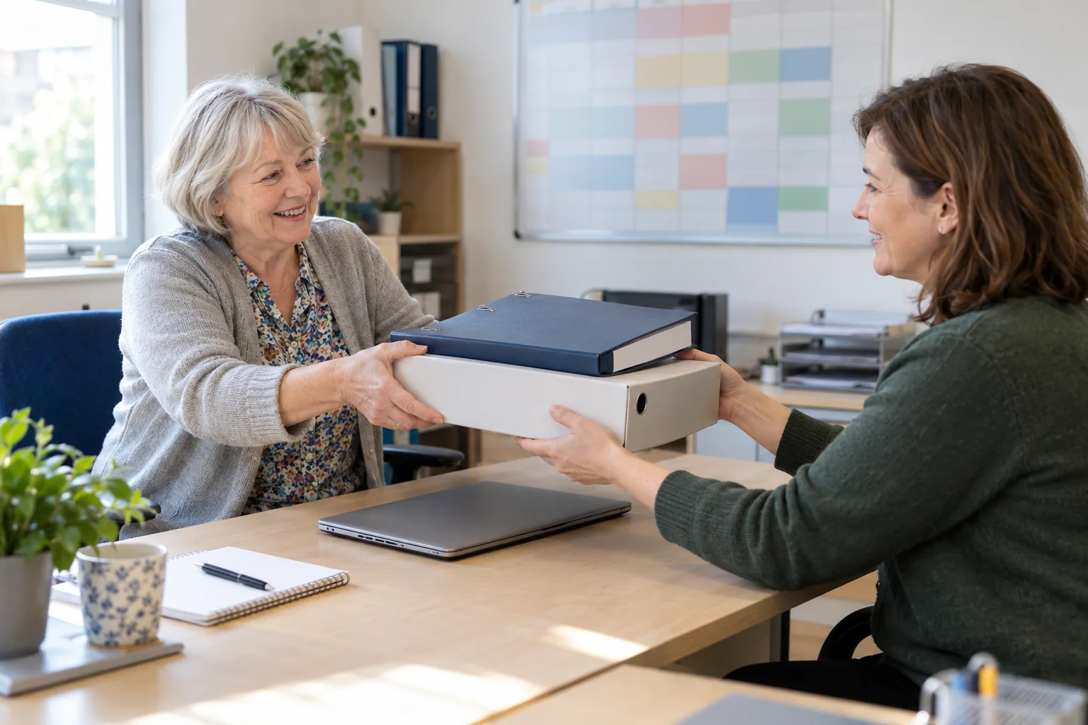 On a bright weekday morning in the small back office of a community centre, an outgoing secretary hands a ring binder and a box file across a desk to the trustee taking over, a laptop closed between them and a wall planner out of focus behind