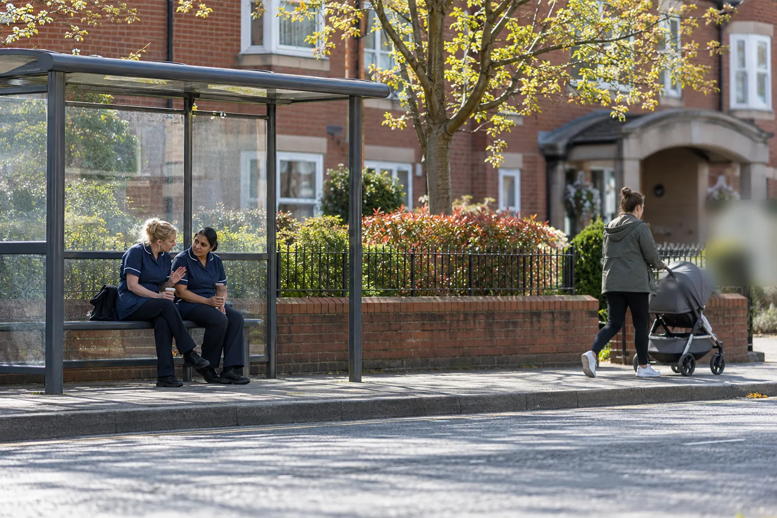 Two care workers in tunics talk quietly on the bench of a bus shelter outside a care home, one lowering her voice as a passer-by walks past with a pushchair