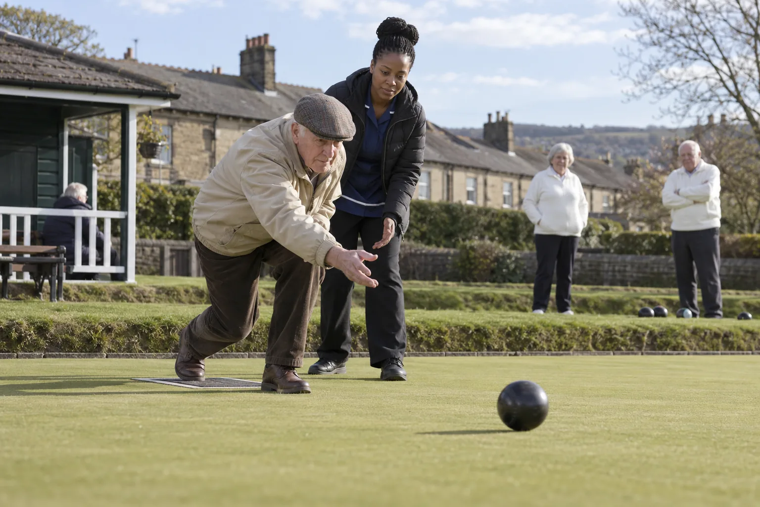 A care home resident in a flat cap bowls on a crown green on a bright afternoon while his key worker stands beside him and two club members look on from the bank