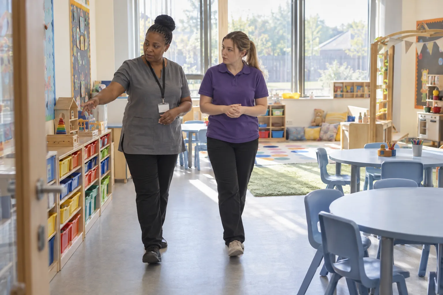 A nursery manager walks a new member of staff through an empty playroom before opening, morning light on the low tables