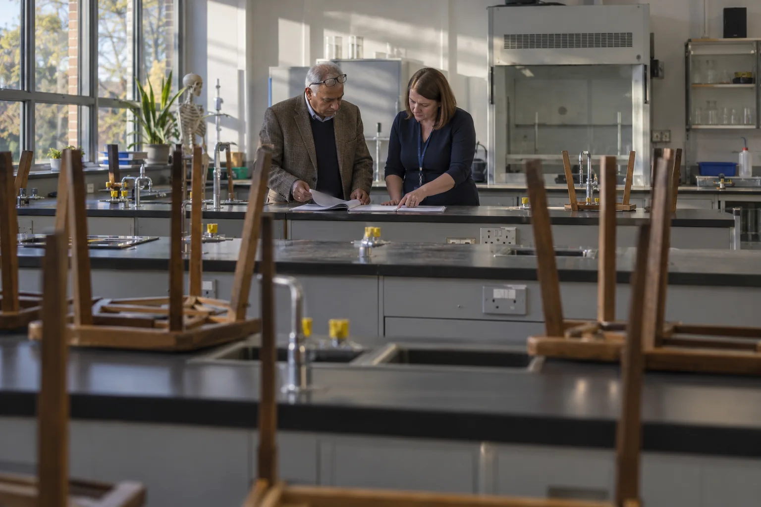 A school governor and the designated safeguarding lead go through a printed annual review at a bench in an empty secondary school science laboratory, stools upturned on the benches around them and late afternoon sun through the windows