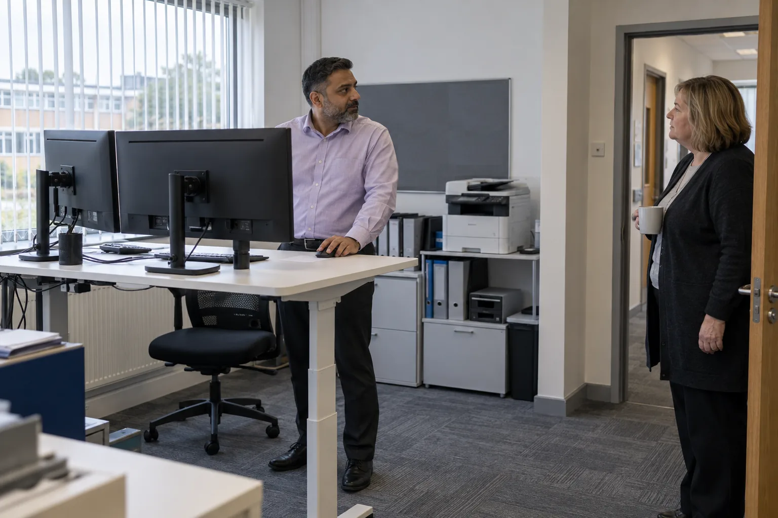 A school data manager at a standing desk with two monitors turned away, a colleague pausing at the office door