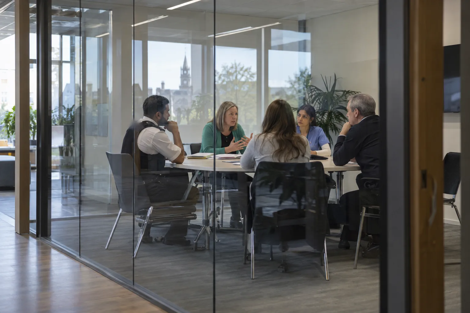 A multi-agency strategy meeting in a council meeting room seen through a glass wall, a police officer, a nurse and three colleagues around a table with paper files