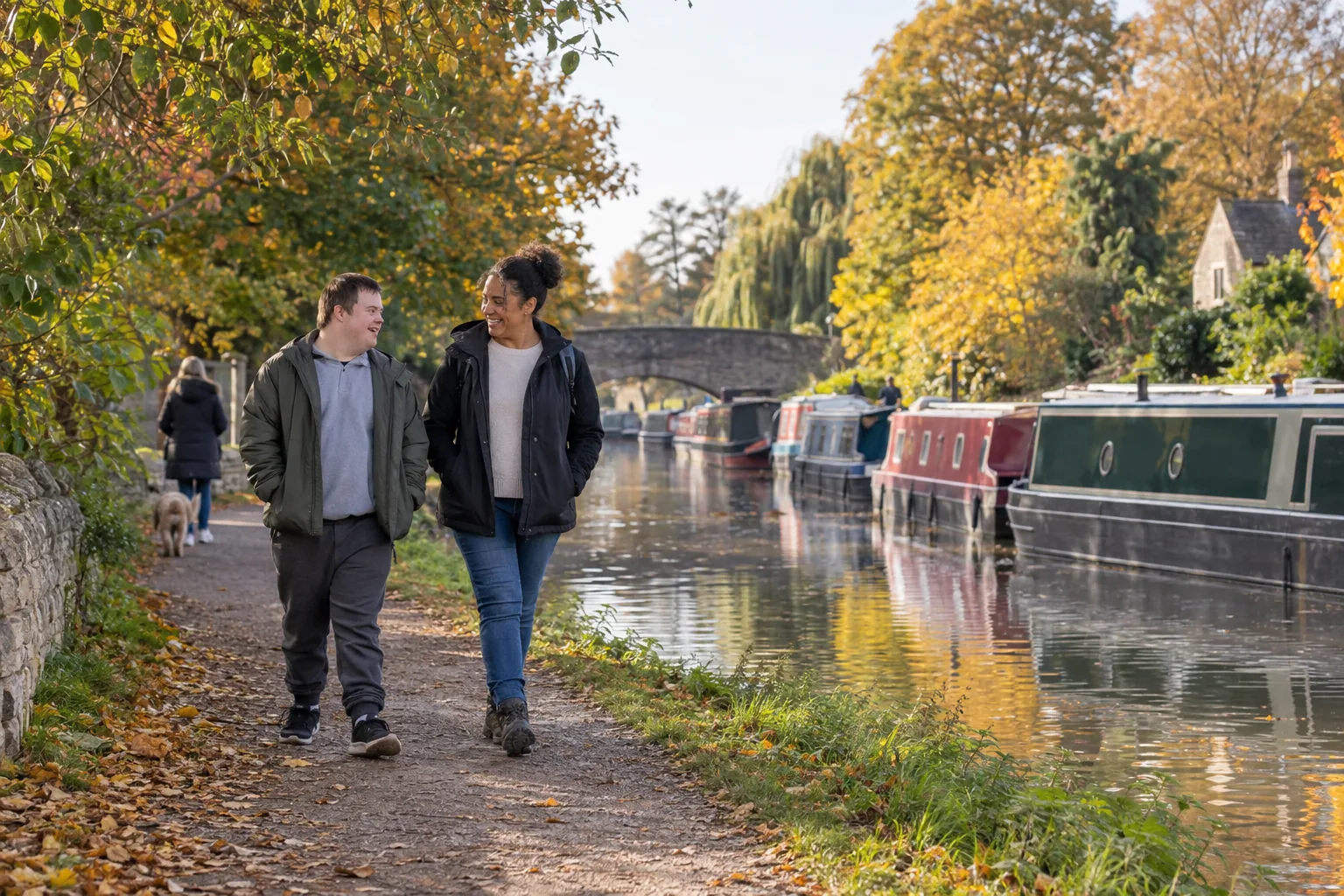 A support worker and a young man with a learning disability walk side by side along a canal towpath on a bright autumn afternoon, talking