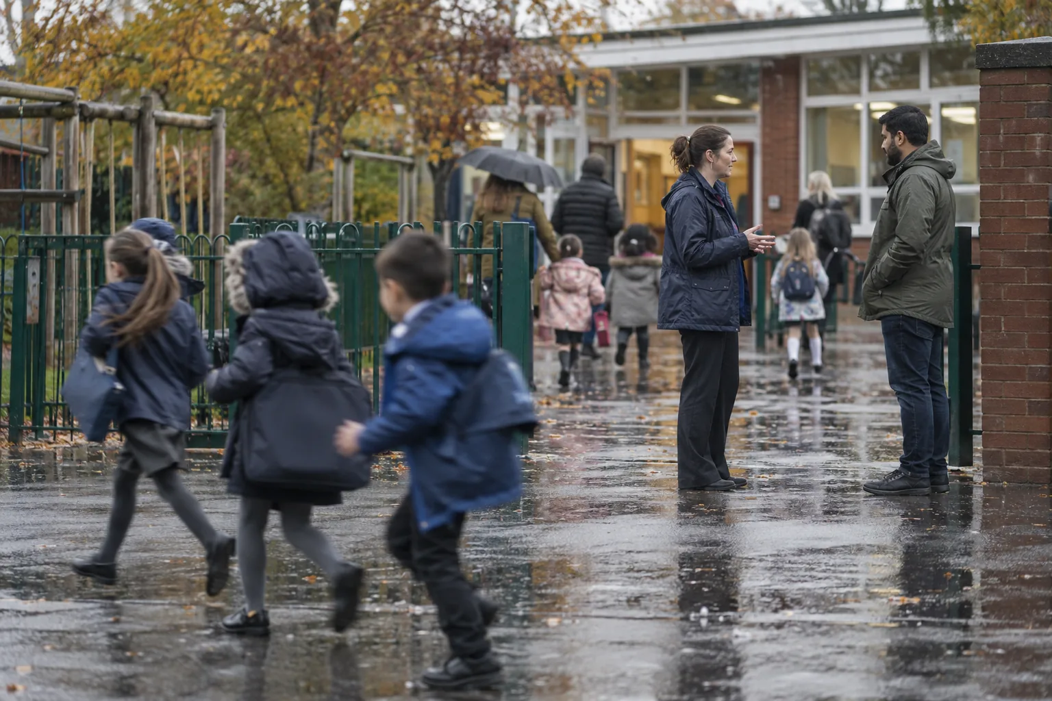 Seen from across a wet primary school playground at drop-off, a deputy head in a rain jacket talks quietly with a parent by the gate while children run past with book bags