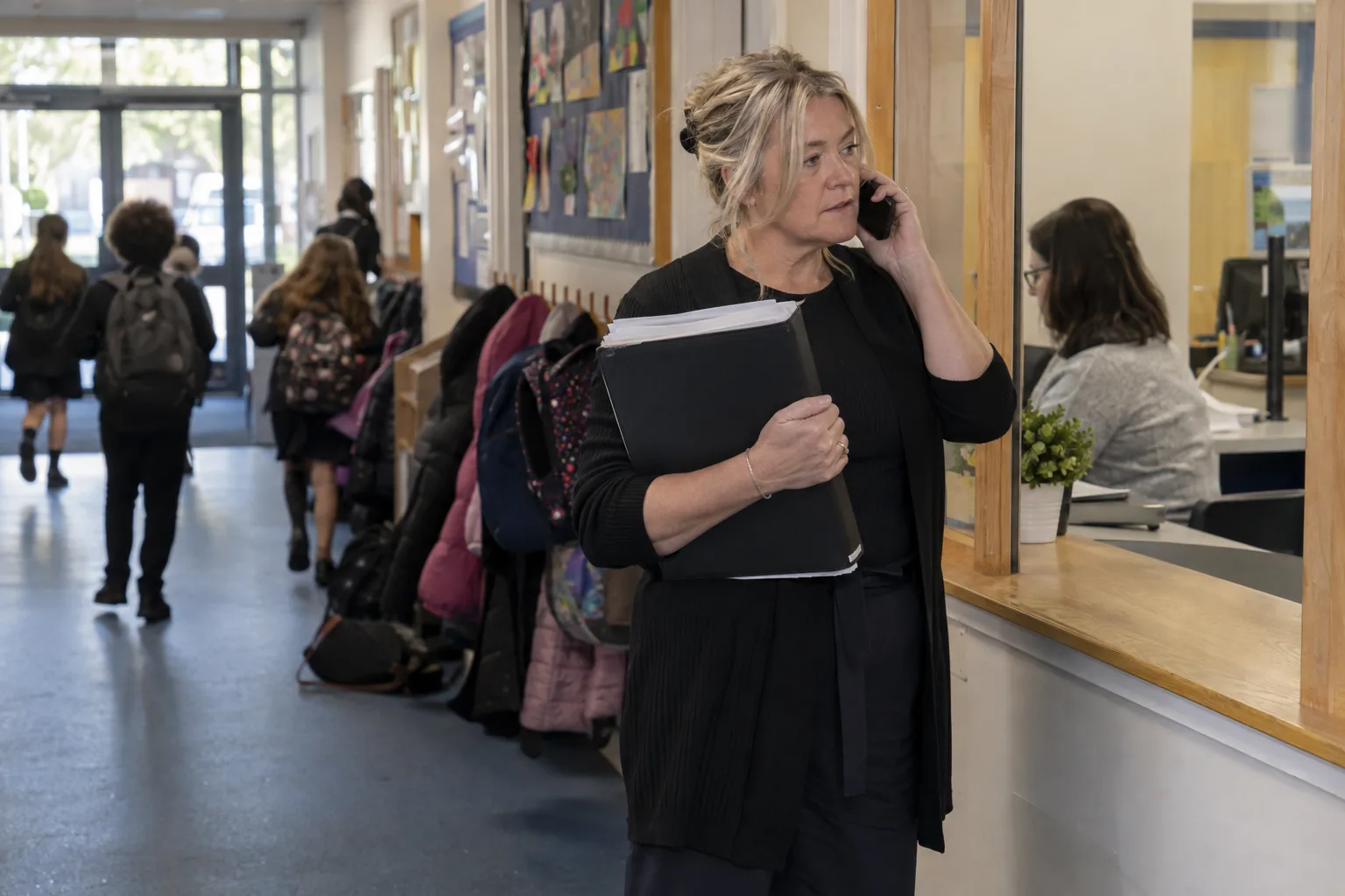 A school business manager stands in the corridor outside the school office late on a Friday afternoon, phone to her ear, the reception hatch and a row of pegged coats behind her