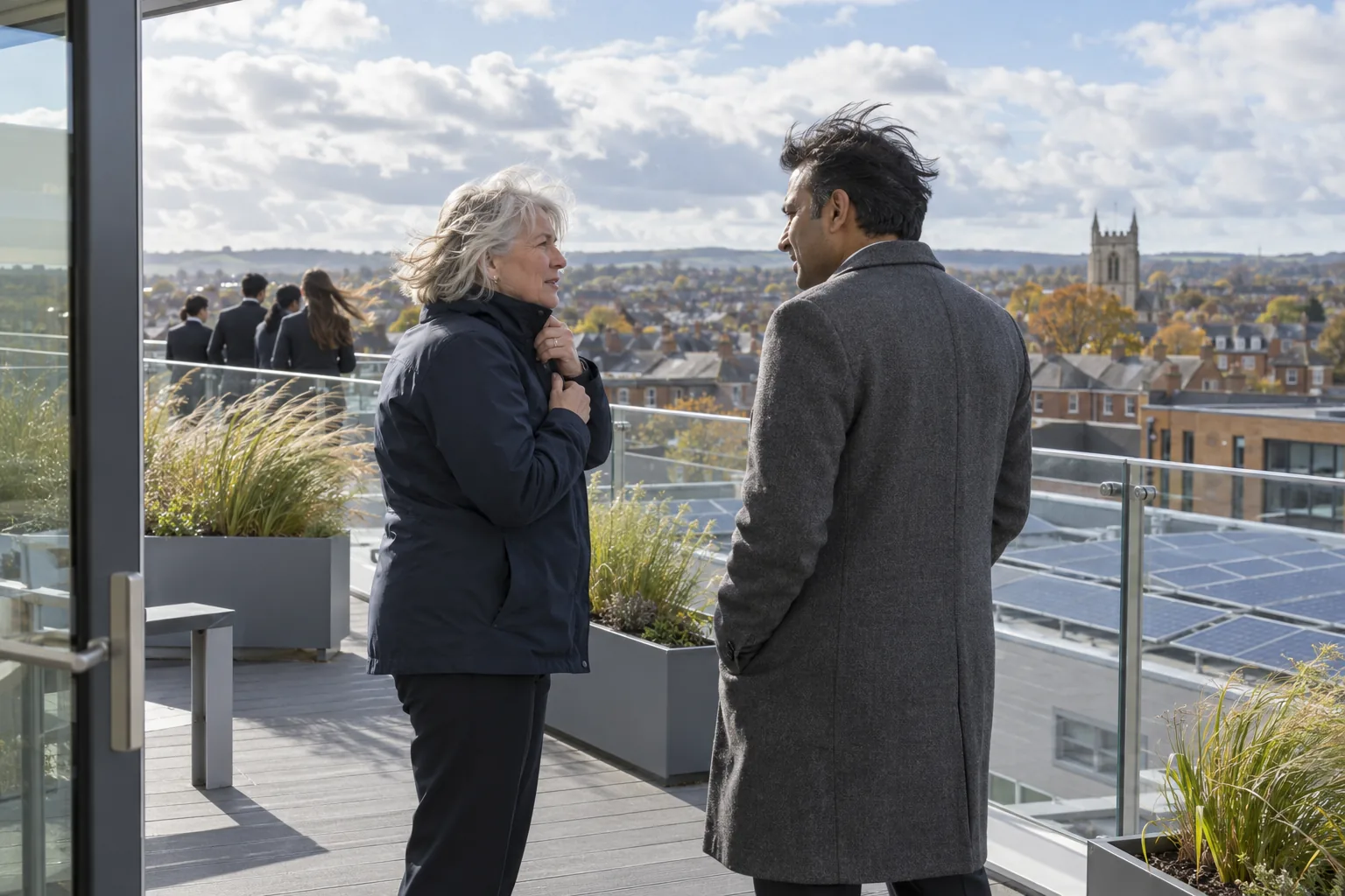 Two academy trustees talk on the open roof terrace of a new-build secondary academy on a bright, windy autumn morning, one zipping up her jacket, the town’s rooftops behind them