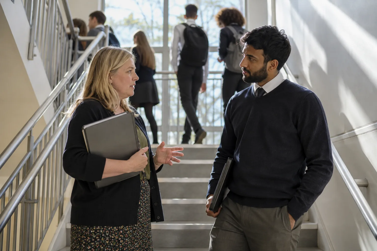 A designated safeguarding lead and a supply teacher talking quietly in a sunlit secondary school stairwell between lessons, pupils blurred passing on the landing above
