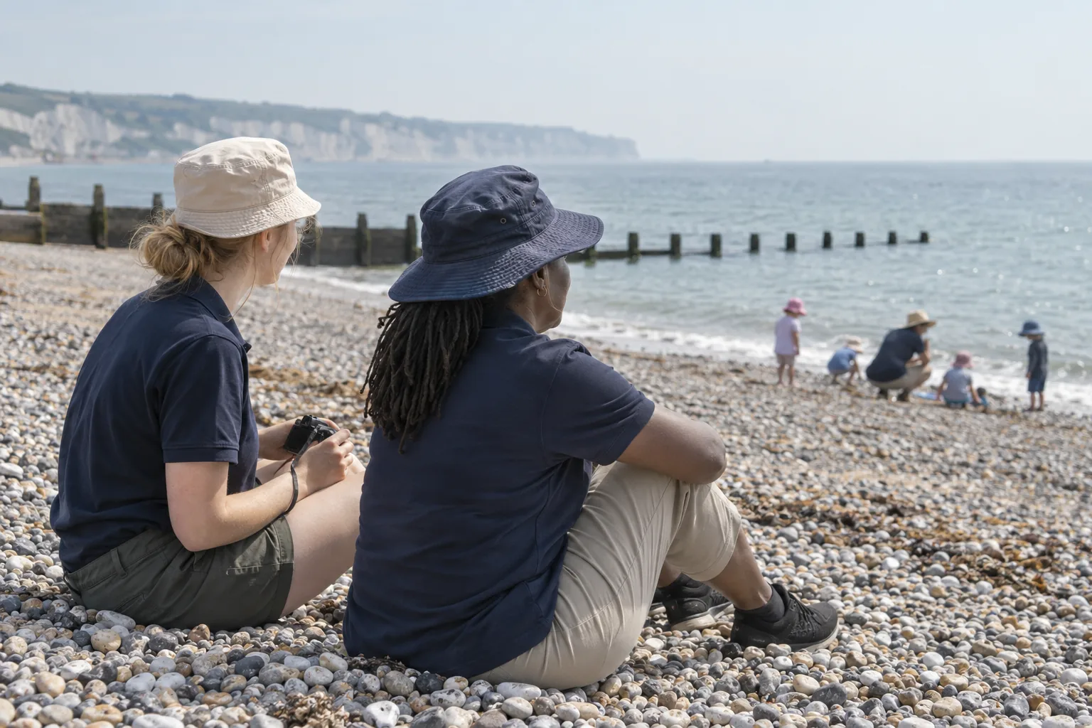 On a nursery’s summer outing to a pebble beach, two practitioners sit on the stones keeping watch, one holding a plain camera in her lap, while small children in sun hats play at the water’s edge in the distance