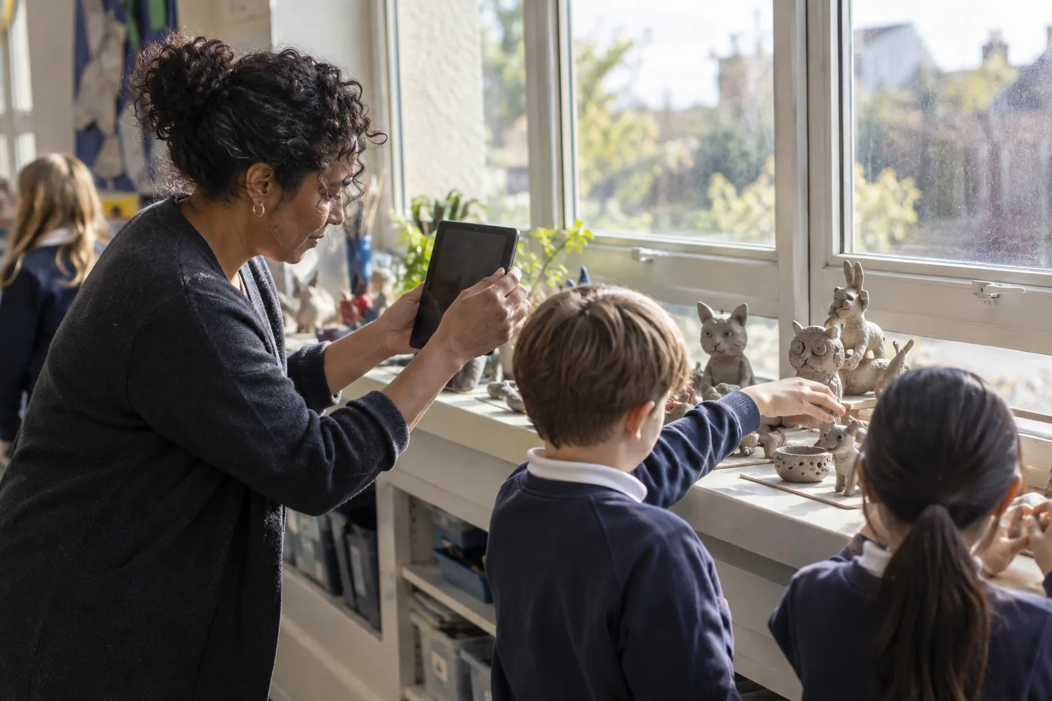 A teaching assistant photographs pupils' clay sculptures laid out on a classroom windowsill with a school-issued tablet, children's hands still arranging the pieces, afternoon light through the window