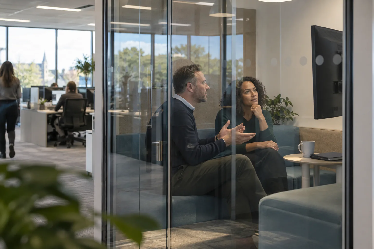Seen from outside through the glass, two staff at a multi-academy trust’s central office talk on a bench seat in a small meeting pod, a wall screen beside them
