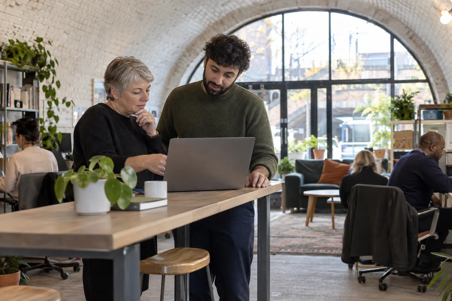 A charity’s operations manager and a volunteer IT adviser talk over a laptop at a high bench in a railway-arch office, the brick vault curving overhead